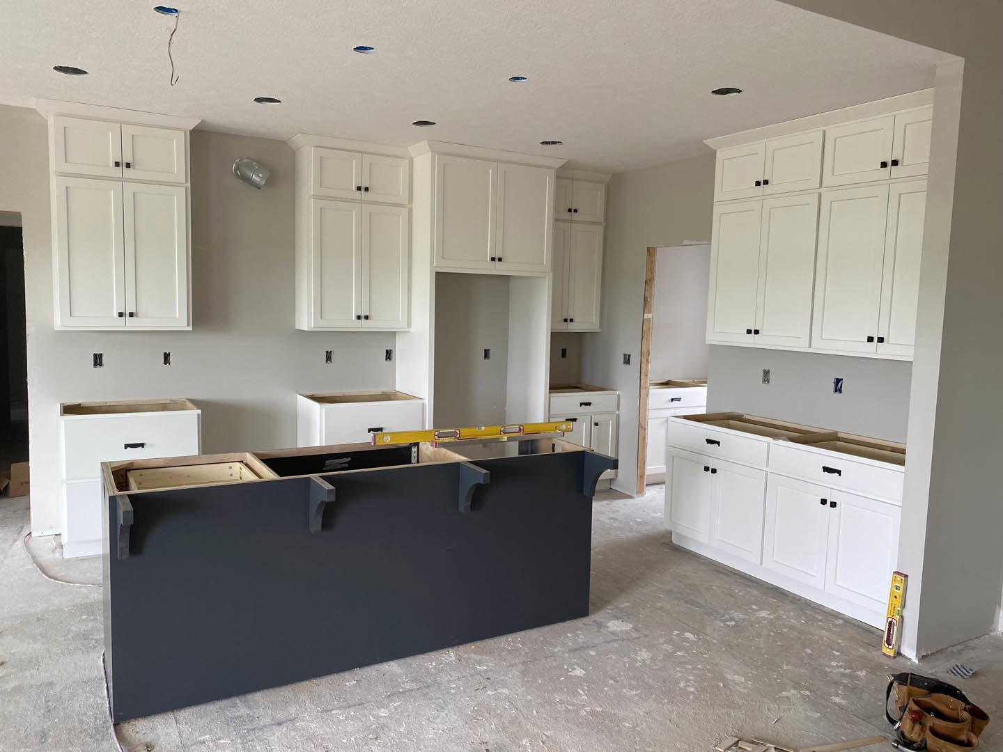 White kitchen cabinets with black handles, black island with matching countertop, yellow level on the counter, brown tool belt on the floor, silver pipe resting on a white surface