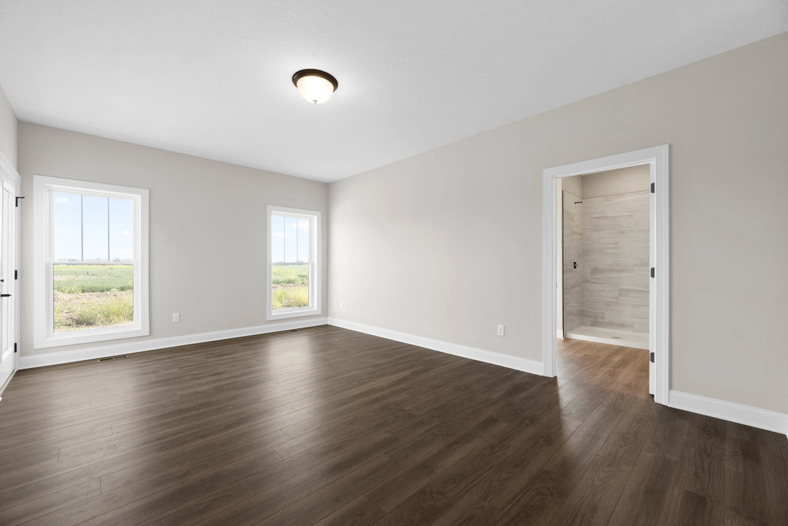 Wood flooring in a spacious room with a ceiling light fixture, white plaster walls, a window overlooking a grassy field, and a glass shower door visible in the background.