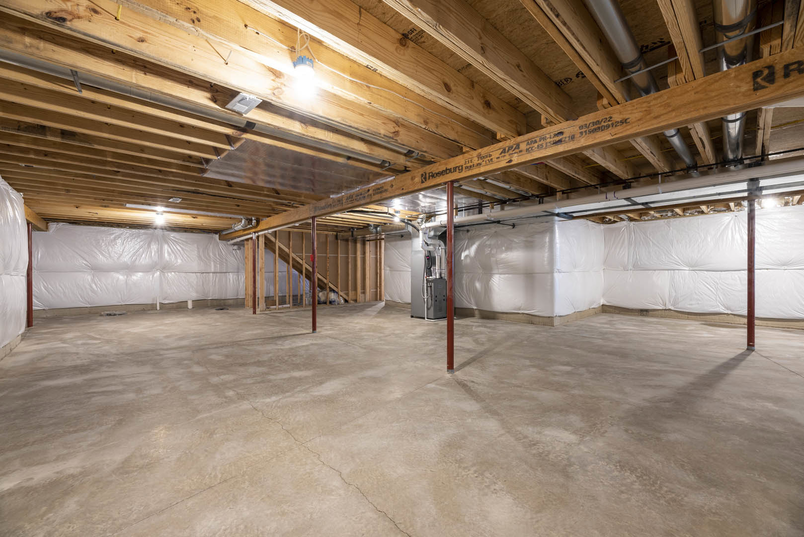 Basement room with exposed wooden ceiling beams, white insulated wall, concrete floor with visible crack, and ceiling-mounted light fixture