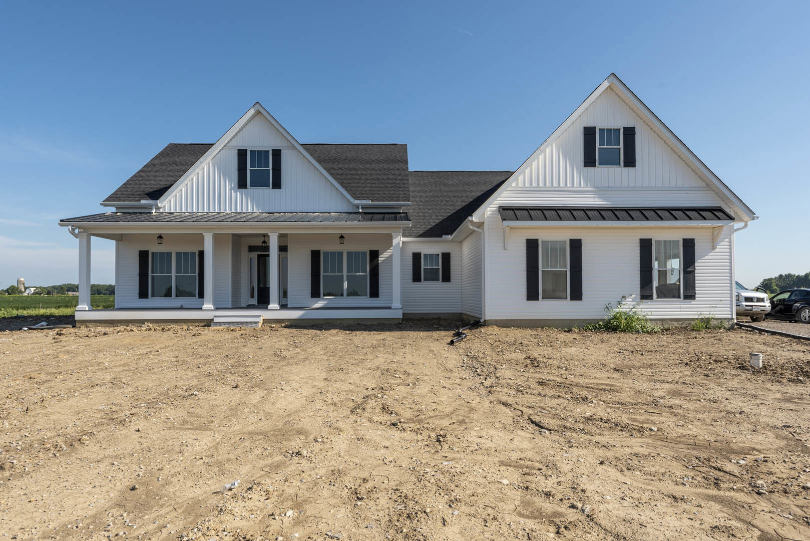 White house with black shutters and white-framed windows, dirt yard with exposed black pipe, open porch, clear sky above