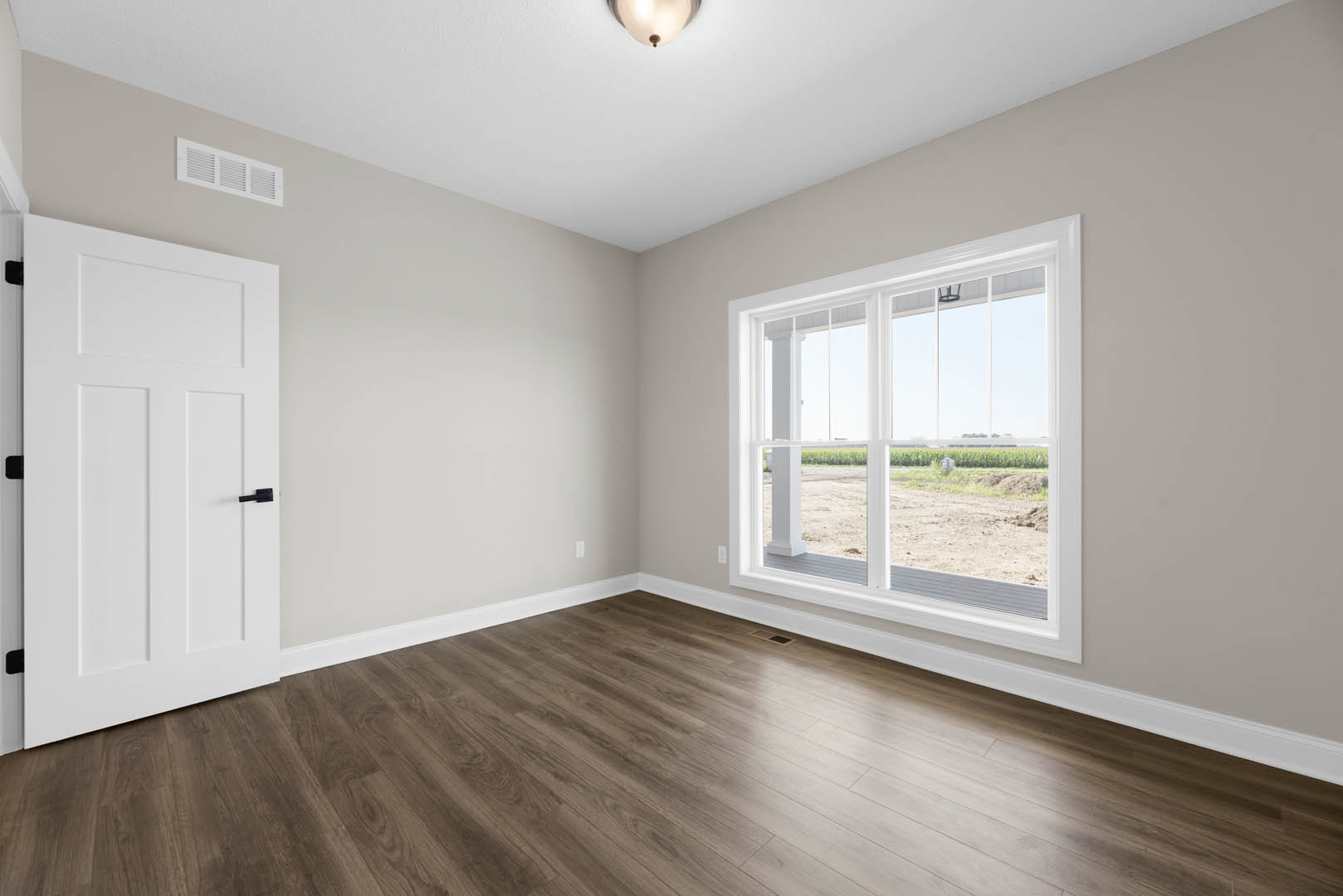 Sunlit room featuring wide-plank hardwood flooring, white walls with base molding, a large window overlooking a grassy field, white door with black handle, wall vent, and recessed