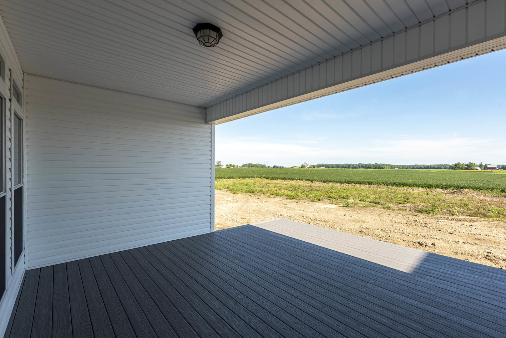 Wood deck with white railing overlooking green field under clear blue sky, outdoor ceiling lamp mounted above, large windows on house exterior