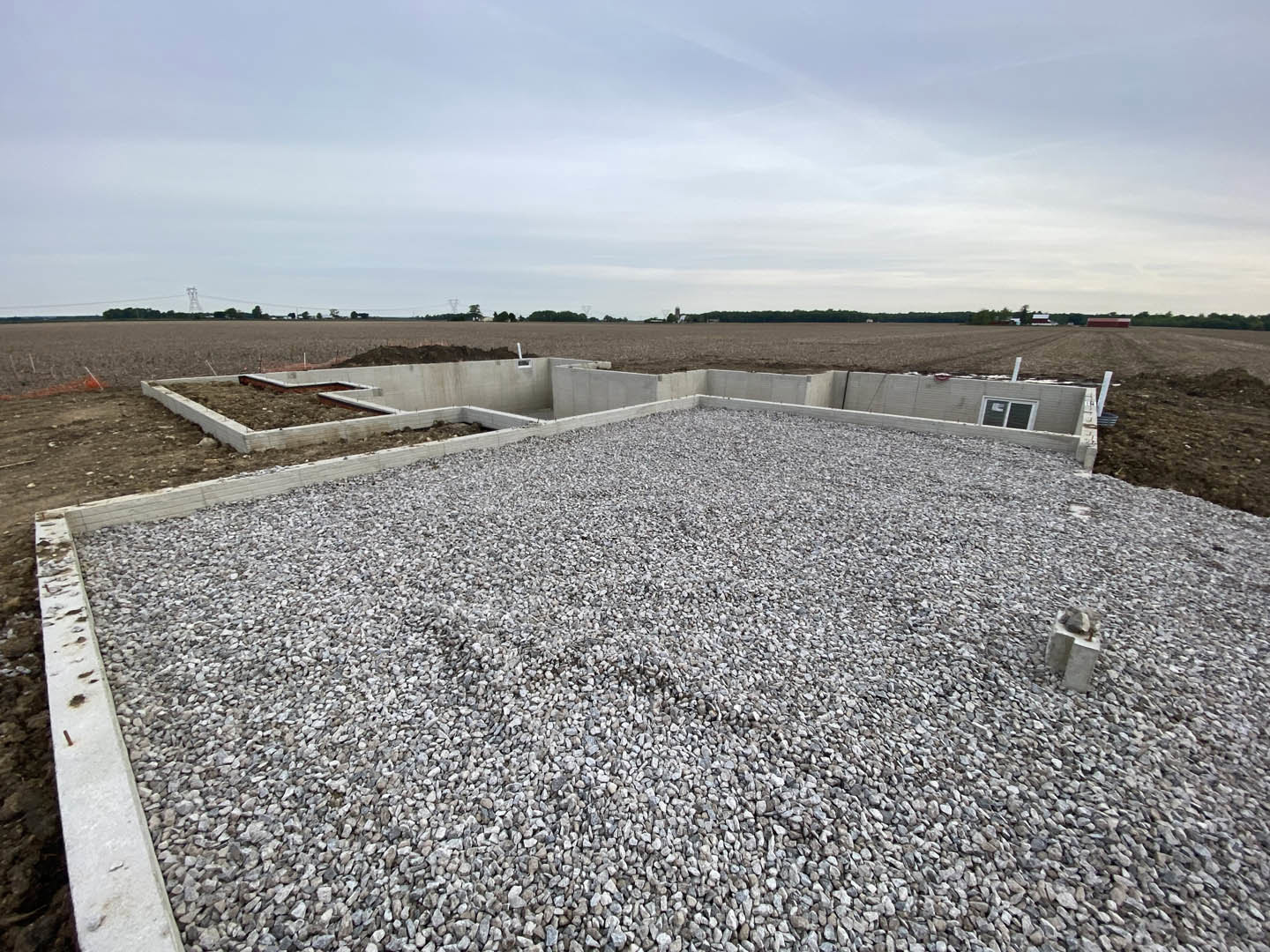 Concrete foundation and curb surrounded by dirt and gravel, construction materials scattered across a large field under a partly cloudy blue sky.