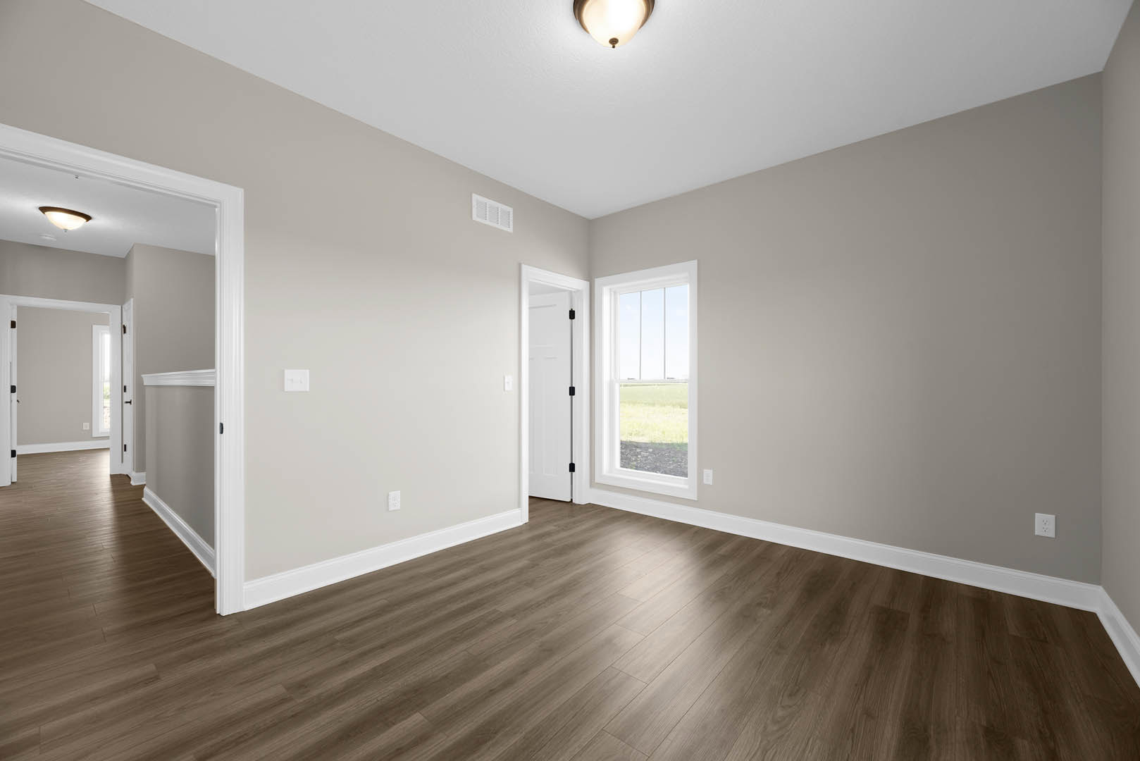 Wood flooring in a bright room with a modern ceiling light fixture, white walls, a window overlooking green grass, and a white door with a black handle.
