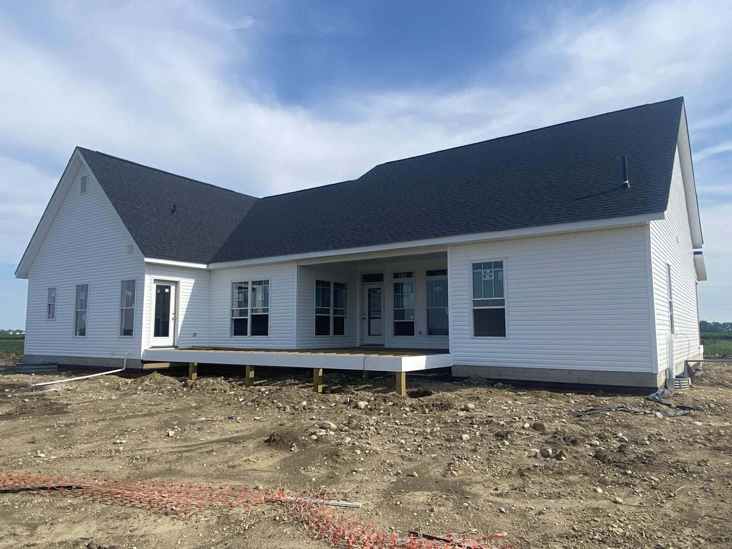 White-framed window set in light siding above a wooden porch platform, dirt ground with construction materials, blue sky and clouds in background