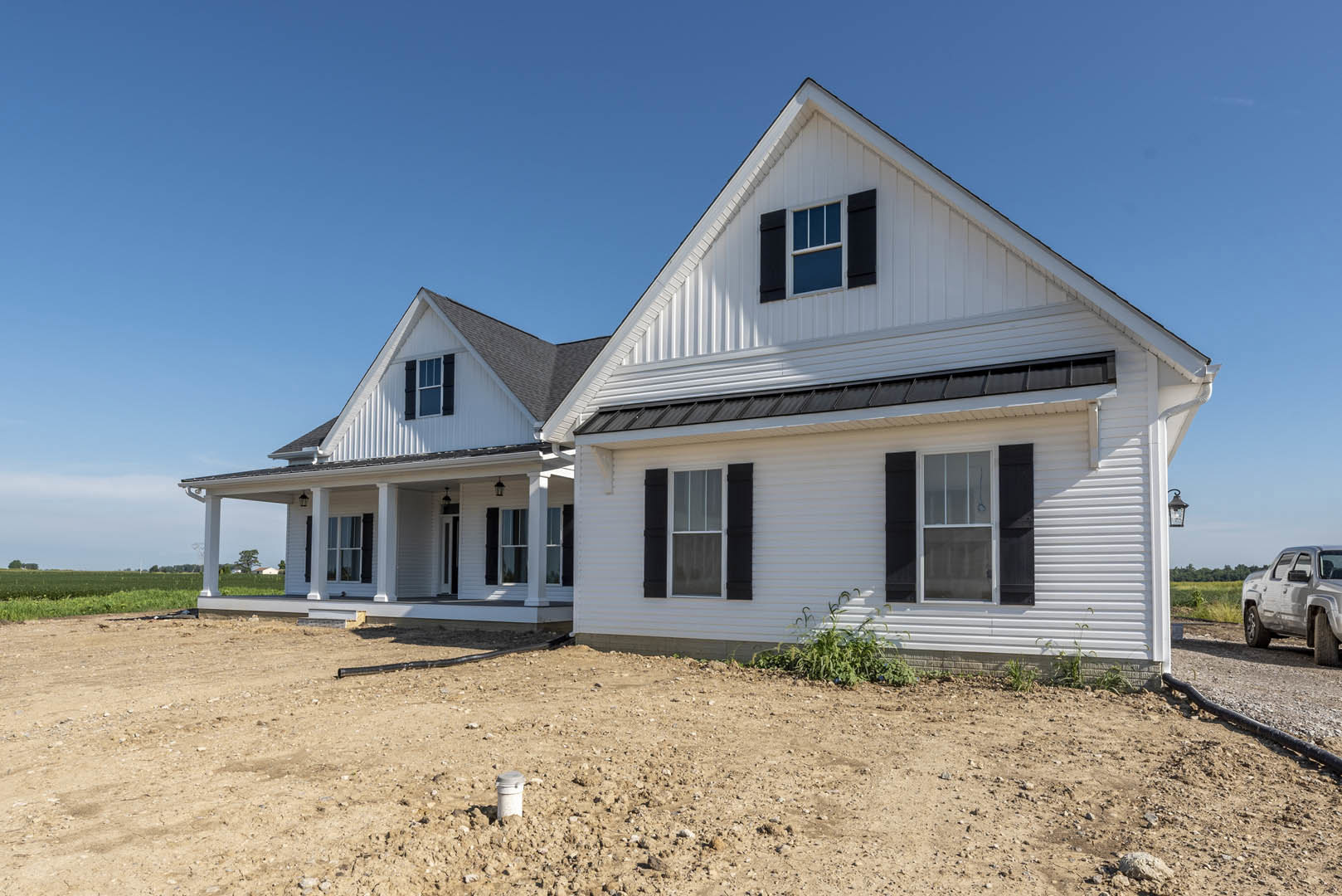 Modern house with black roof and white siding, black shuttered windows, silver truck parked in front, surrounded by a dirt field under a clear sky