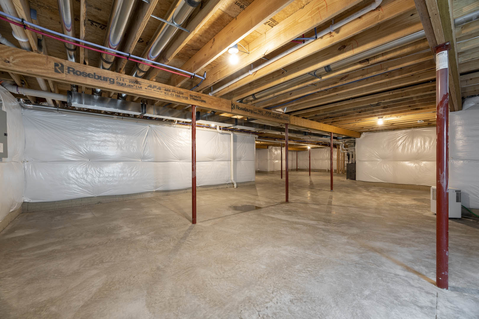 Basement with concrete floor, exposed ceiling beams, white plastic sheeting covering one wall, visible pipes, and illuminated ceiling light fixture