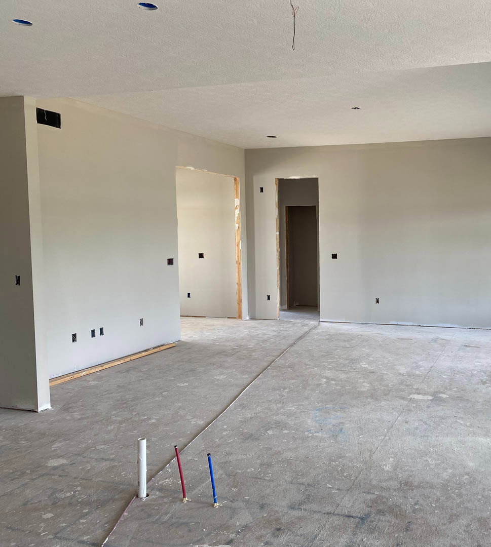 Unfinished room with white plaster walls and ceiling, exposed concrete floor with protruding pipes, brown-framed opening in wall, red and blue poles on floor, minimal construction