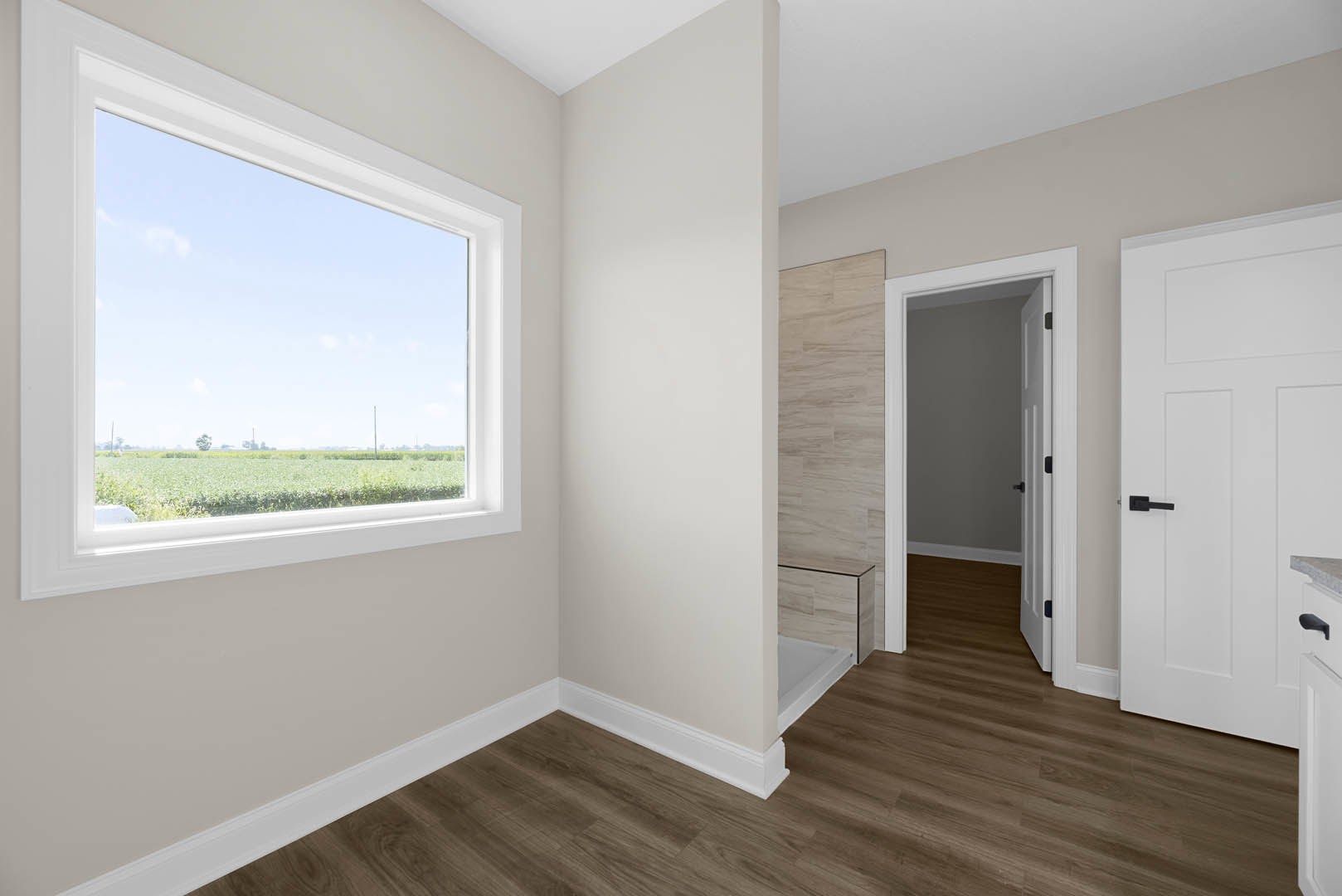 Sunlit room featuring wide wood plank flooring, crisp white walls, large window overlooking green field and bushes, and white door with black handle