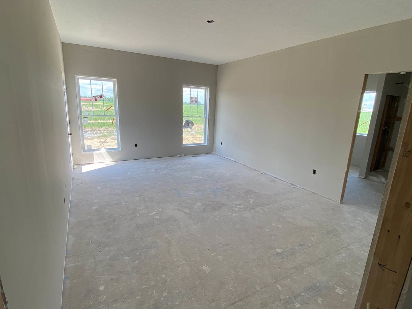 Spacious room featuring light-colored plaster walls, multiple windows with white trim, grey flooring, and a closed door; one window displays a piece of paper taped to the glass.