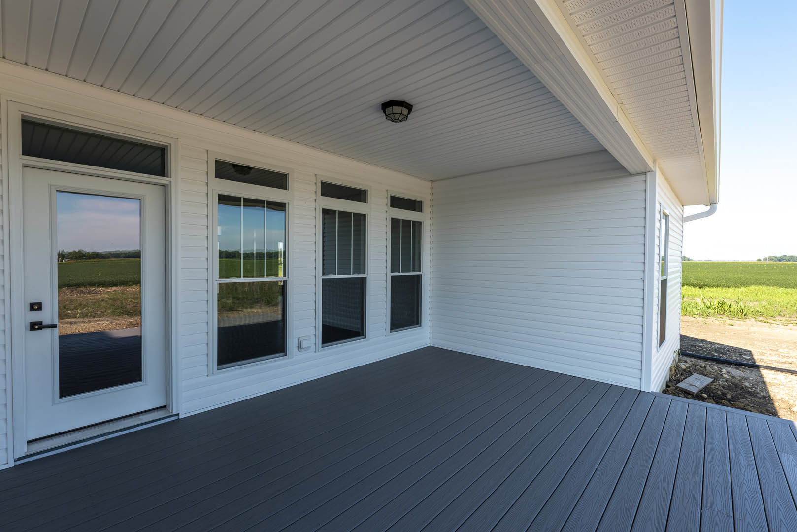 White siding house with large windows, wooden deck, white porch columns, and glass door, set against grassy yard and open sky.