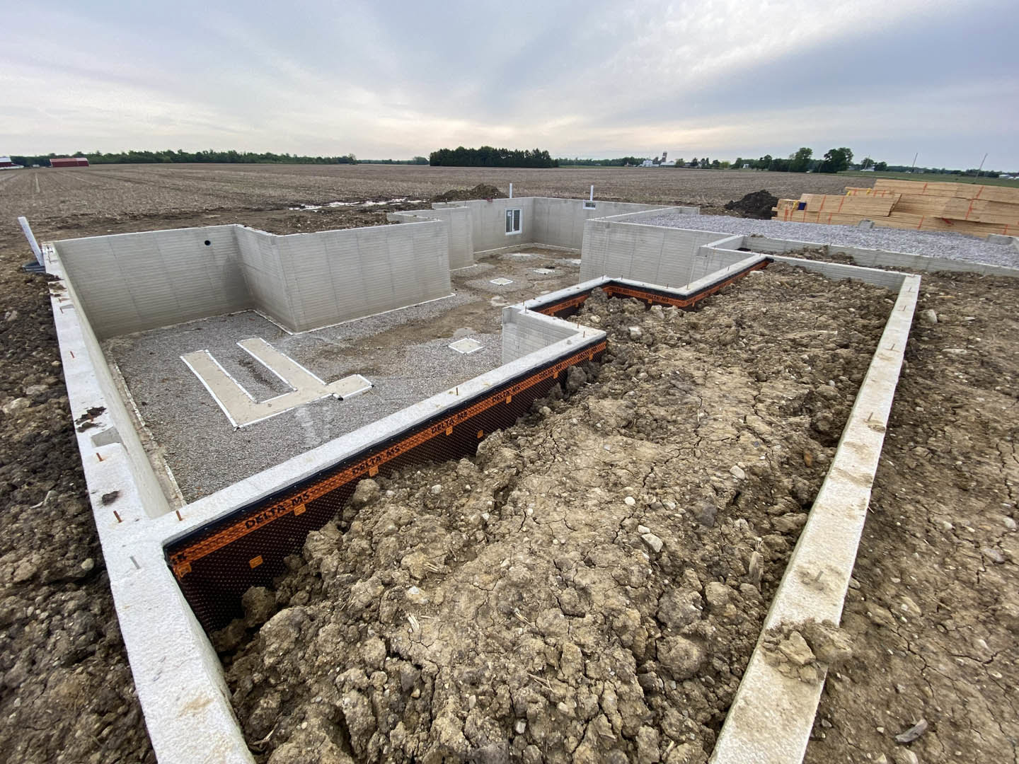 Concrete foundation walls surrounded by dirt and soil, wood planks scattered on the construction site, blue sky with clouds overhead