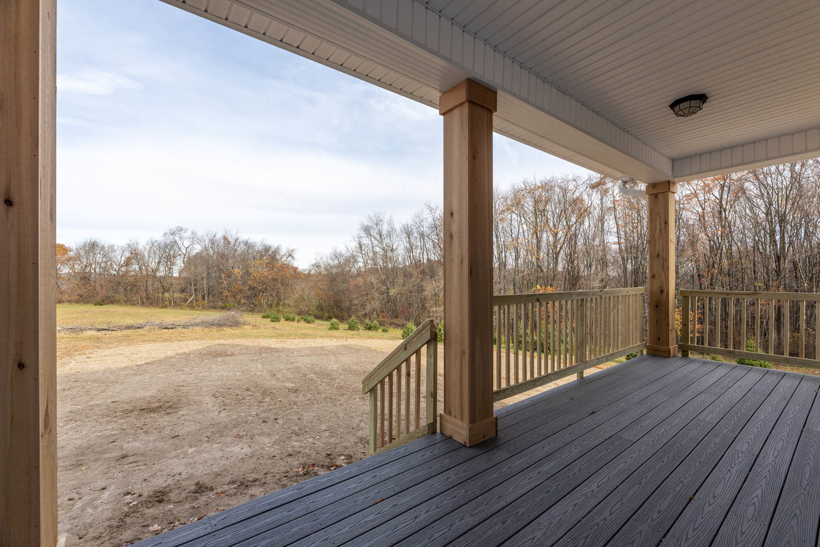 Wooden deck with horizontal railing overlooking a dirt field, trees in the background, light fixture mounted on exterior wall, wooden post featuring a circular hole