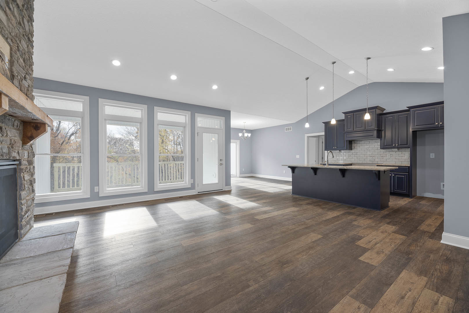 Open kitchen with white cabinetry, large central island, hardwood flooring, white walls, multiple windows overlooking trees, and a white door.