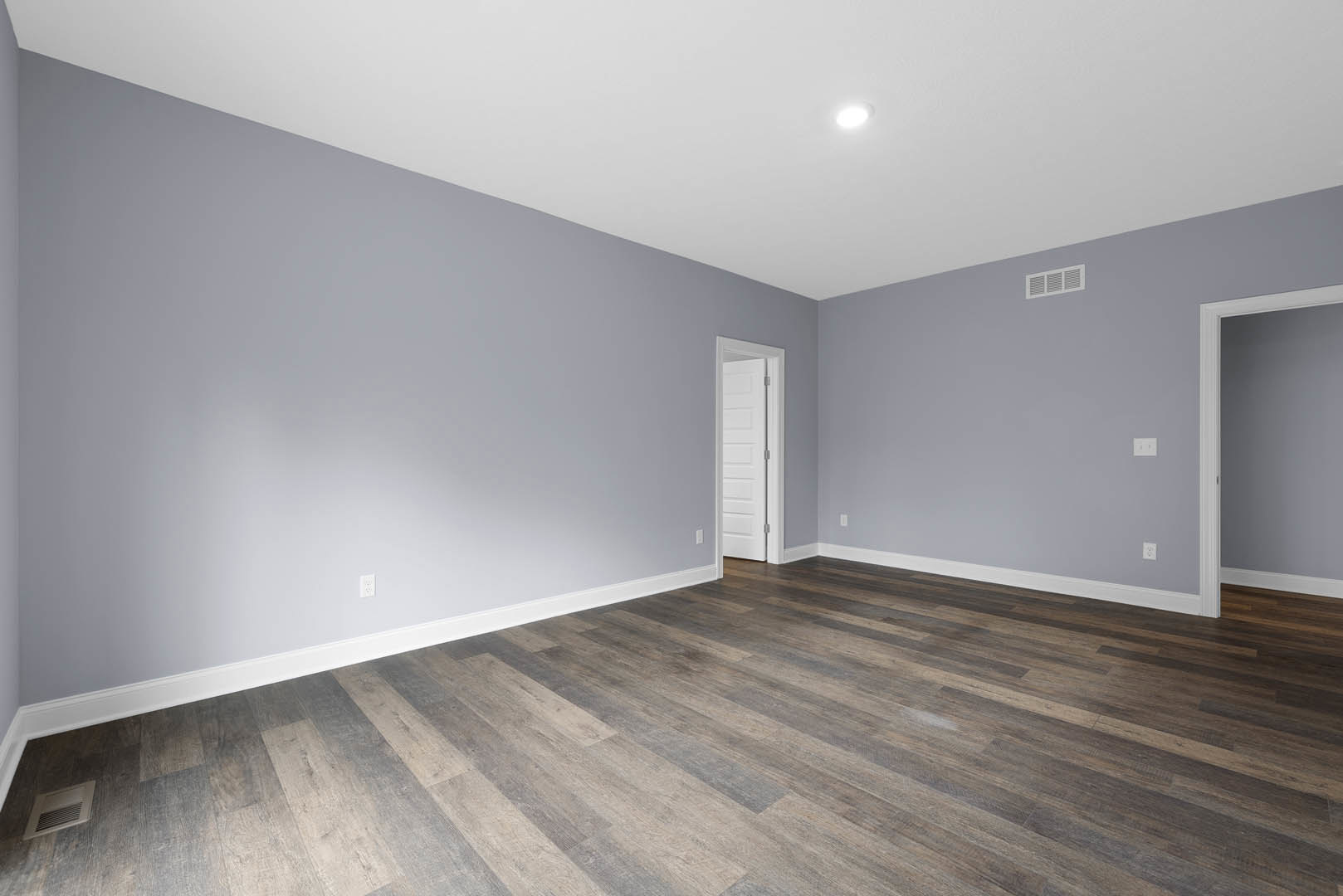 Wood flooring and white walls in a room featuring a white door with a metal handle, ceiling light fixture, and wall vent.