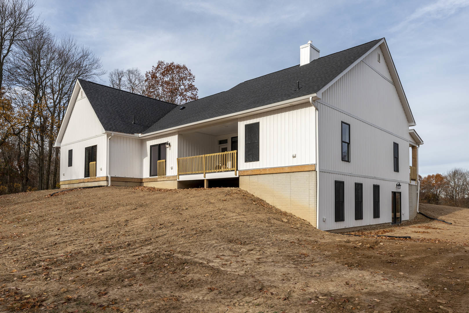 Two-story house with triangular roof and large windows, wooden deck featuring railing with a cat, garage door at ground level, surrounded by grassy hill, Robert Frost Farm visible