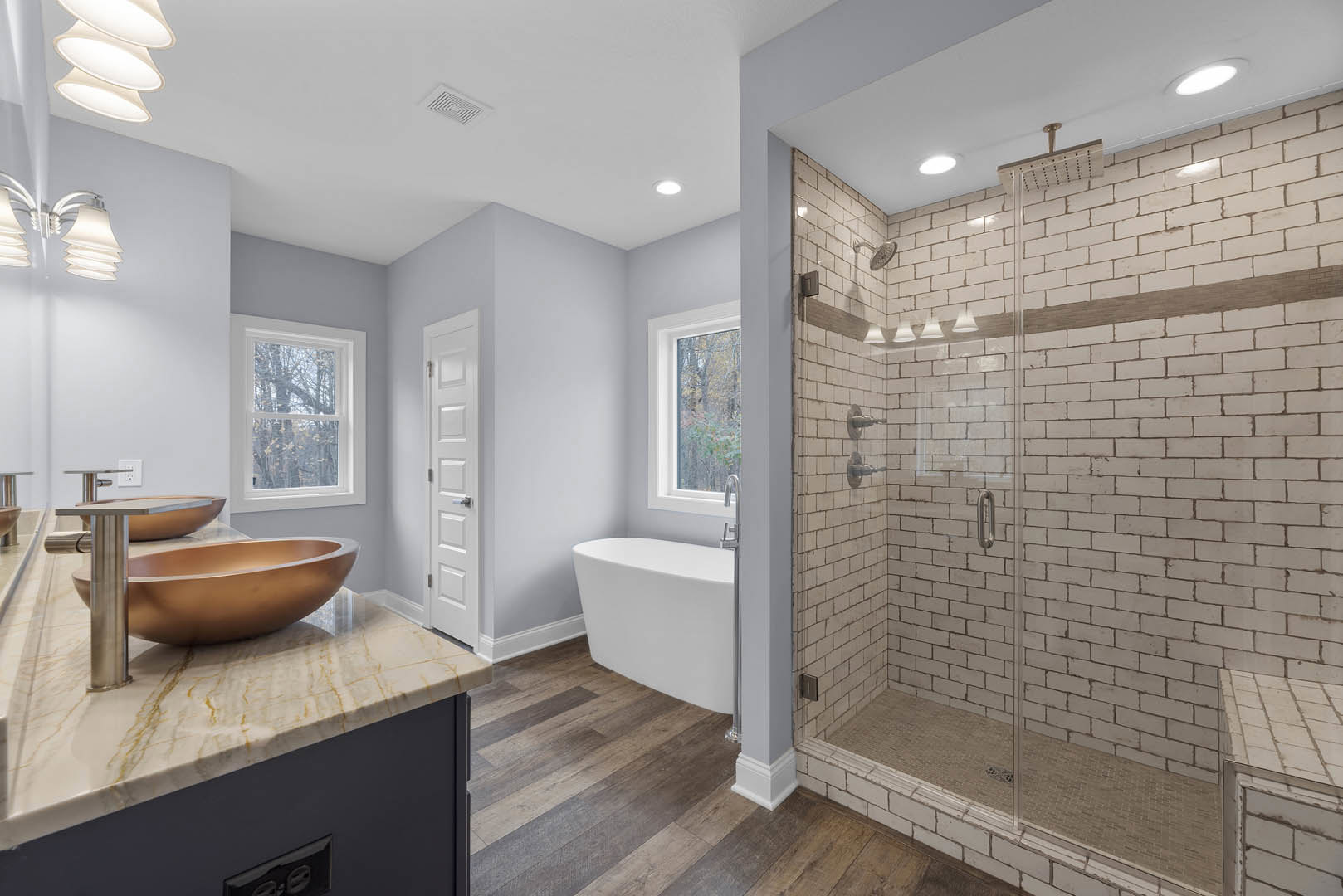 Freestanding white tub beside a marble vanity with a brown vessel sink, glass shower enclosure, cluster of white pendant lamps, large window overlooking trees, light tile flooring