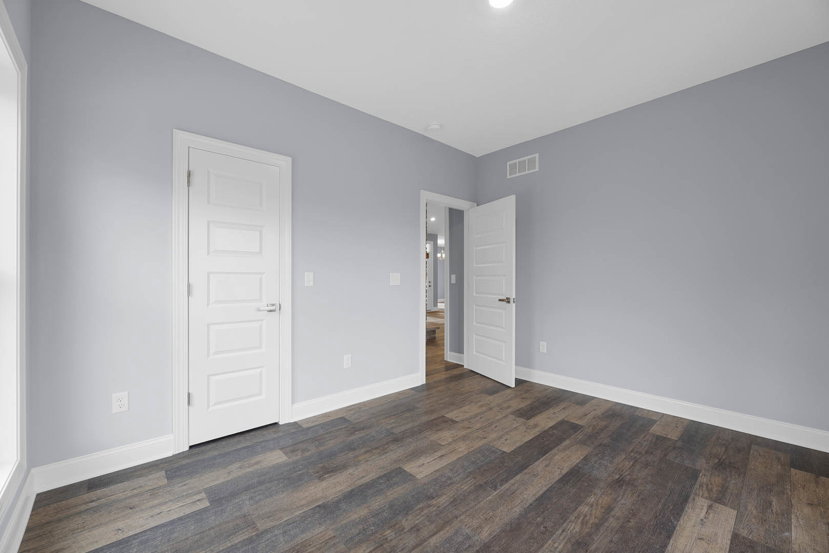White paneled doors with silver handles, wood flooring with white baseboards, wall vent, and white ceiling with recessed light.