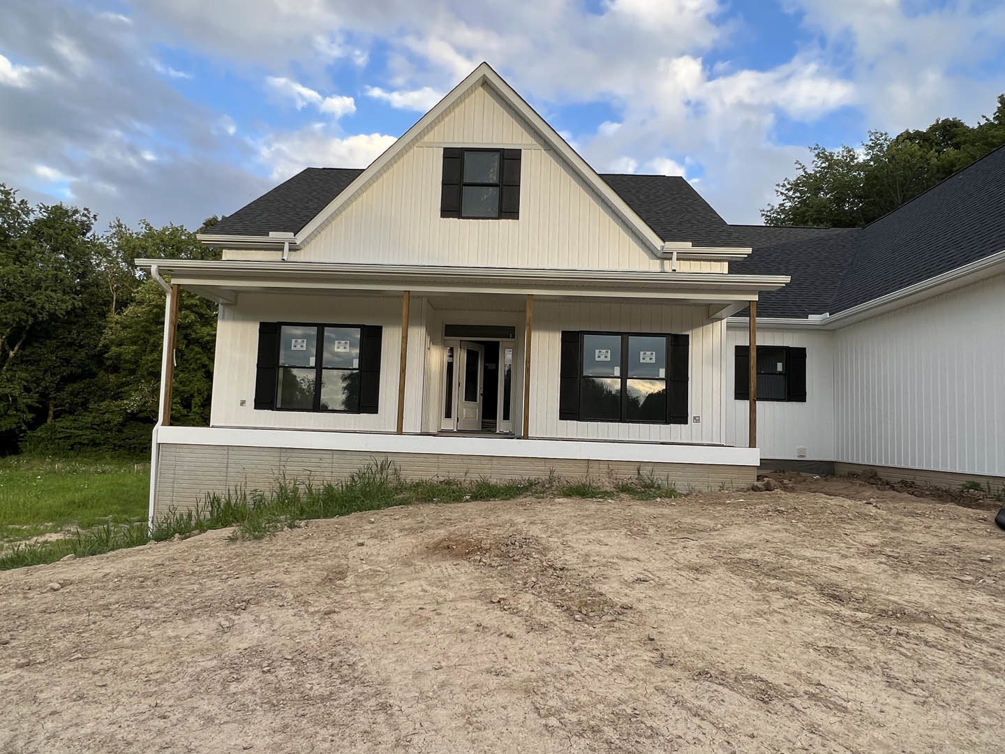 Single-story home with gray siding, black-framed windows displaying stickers, white glass-paneled front door, covered porch, dirt driveway bordered by grass, cloudy sky overhead