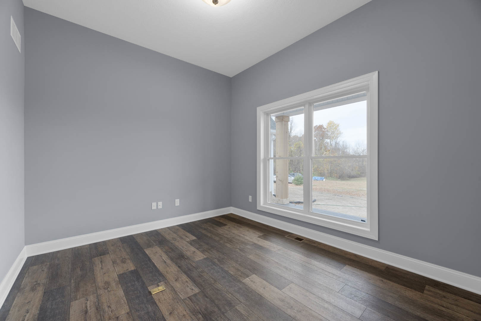 Hardwood floor room with large window overlooking house and trees, white walls, ceiling light fixture, and rectangular wall vent