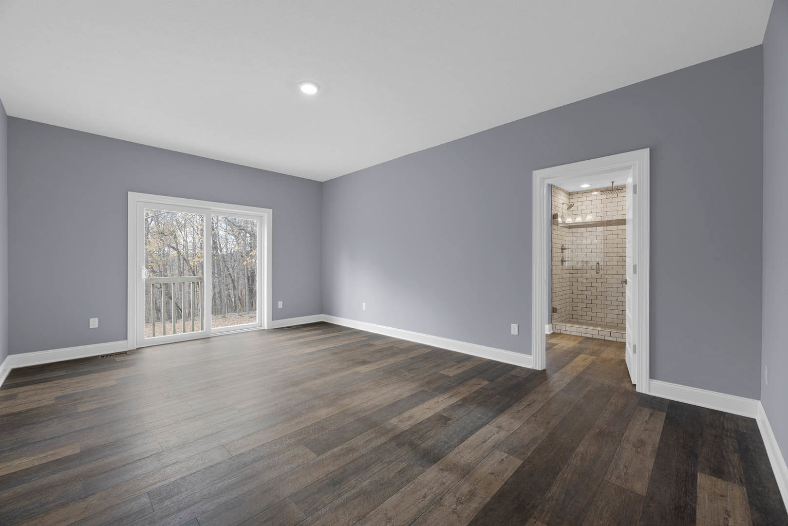 Glass shower enclosure with white subway tile walls, wood flooring with white baseboards, open door leading to view of wooded landscape through sliding glass door