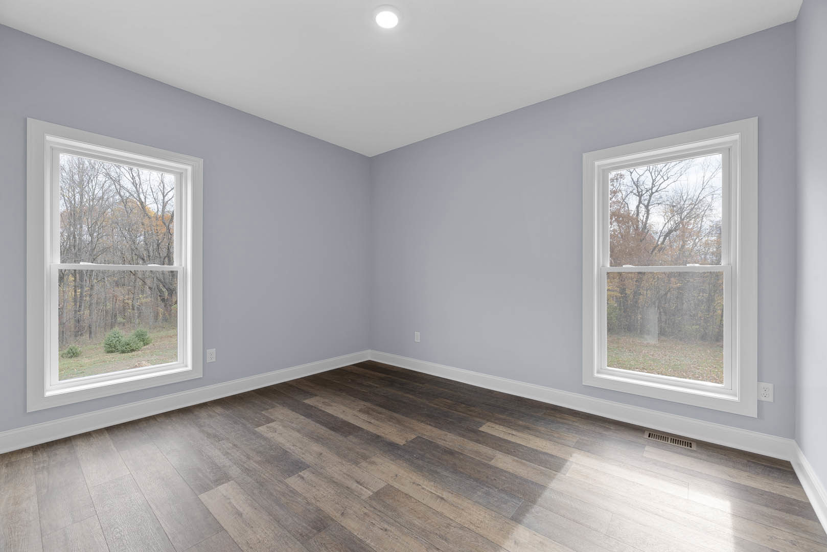 Spacious room with large windows overlooking trees, polished hardwood floor, white plaster walls, and recessed ceiling light