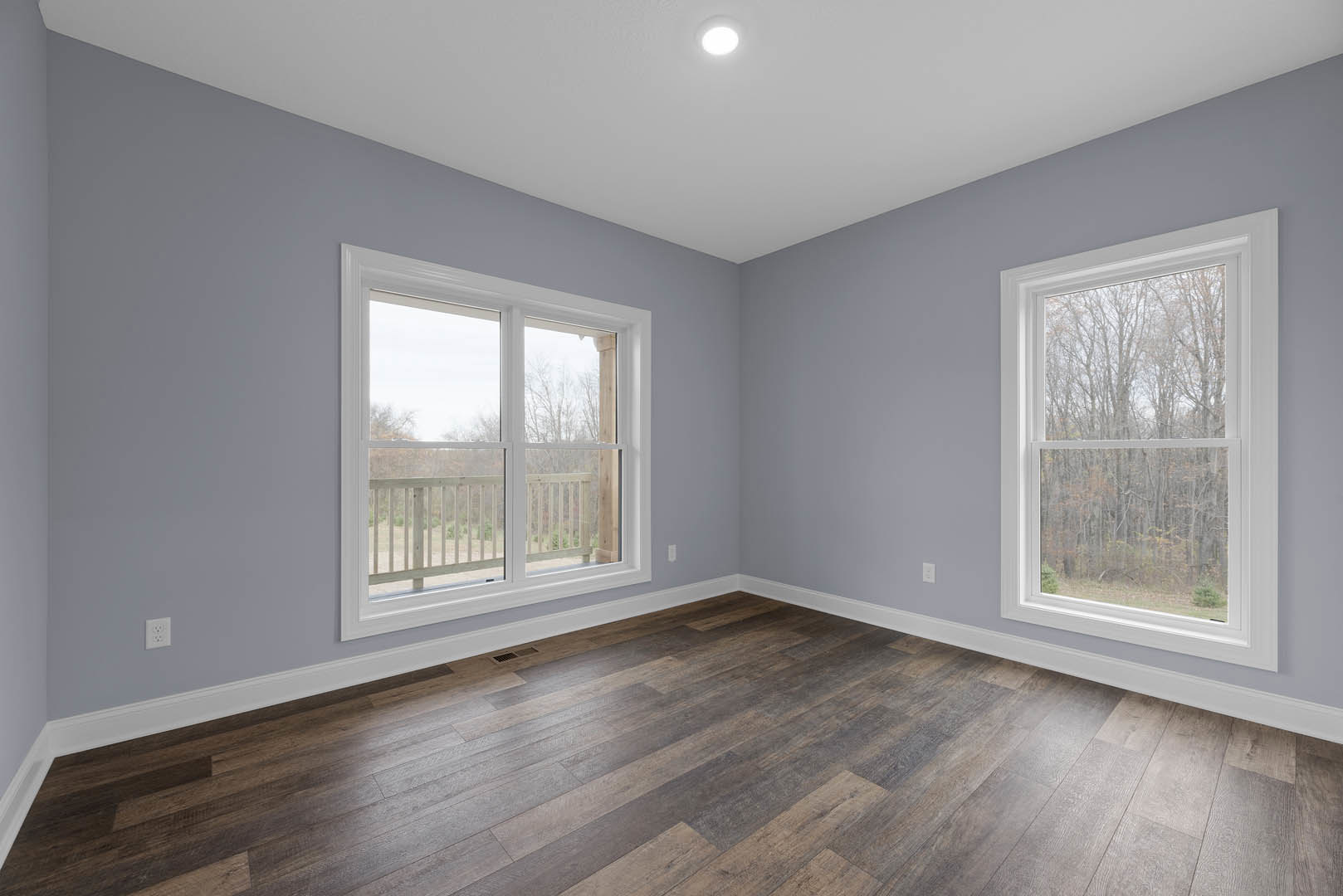 Open room with wide plank wood flooring, large windows overlooking deck and trees, white plaster walls, recessed ceiling light