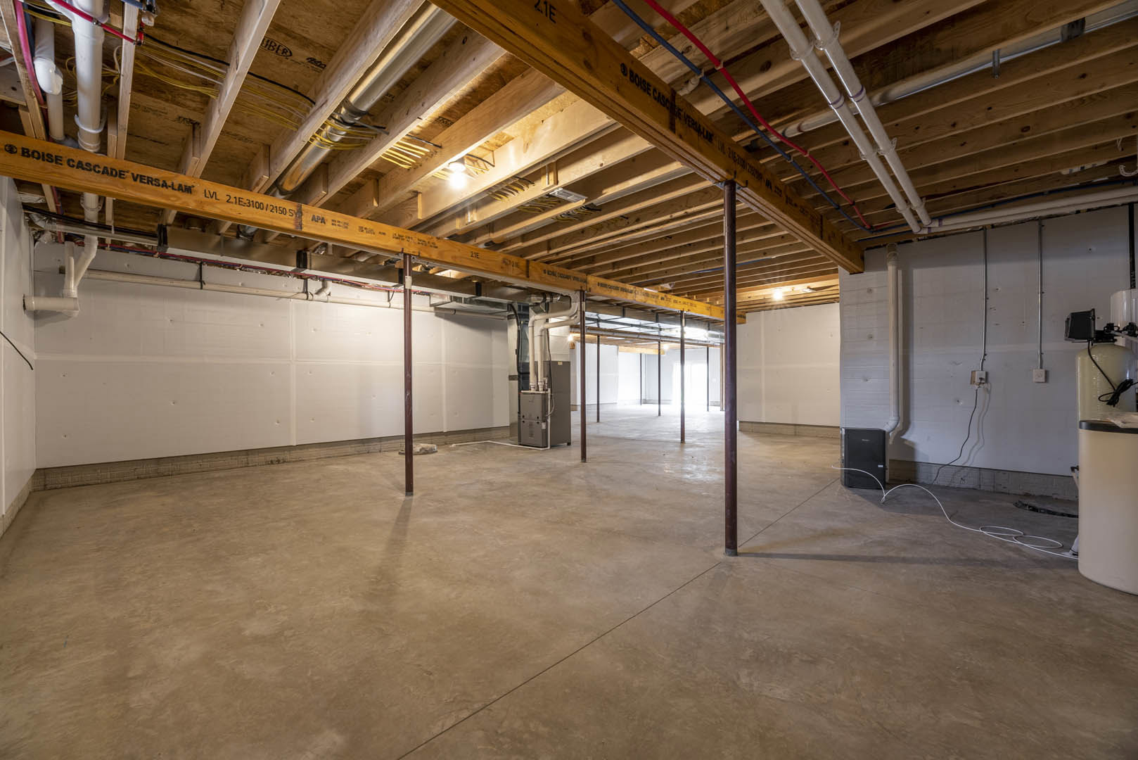 Basement room with exposed wooden ceiling beam, visible pipes, concrete floor, metal support pole, and insulated walls.