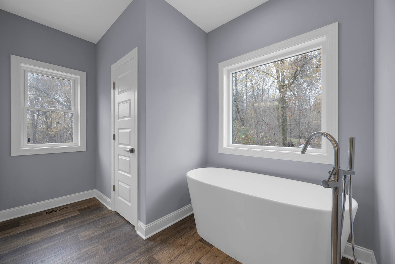 Freestanding white bathtub on wood floor beneath large windows with tree views, plaster walls, chrome faucet, and white door visible