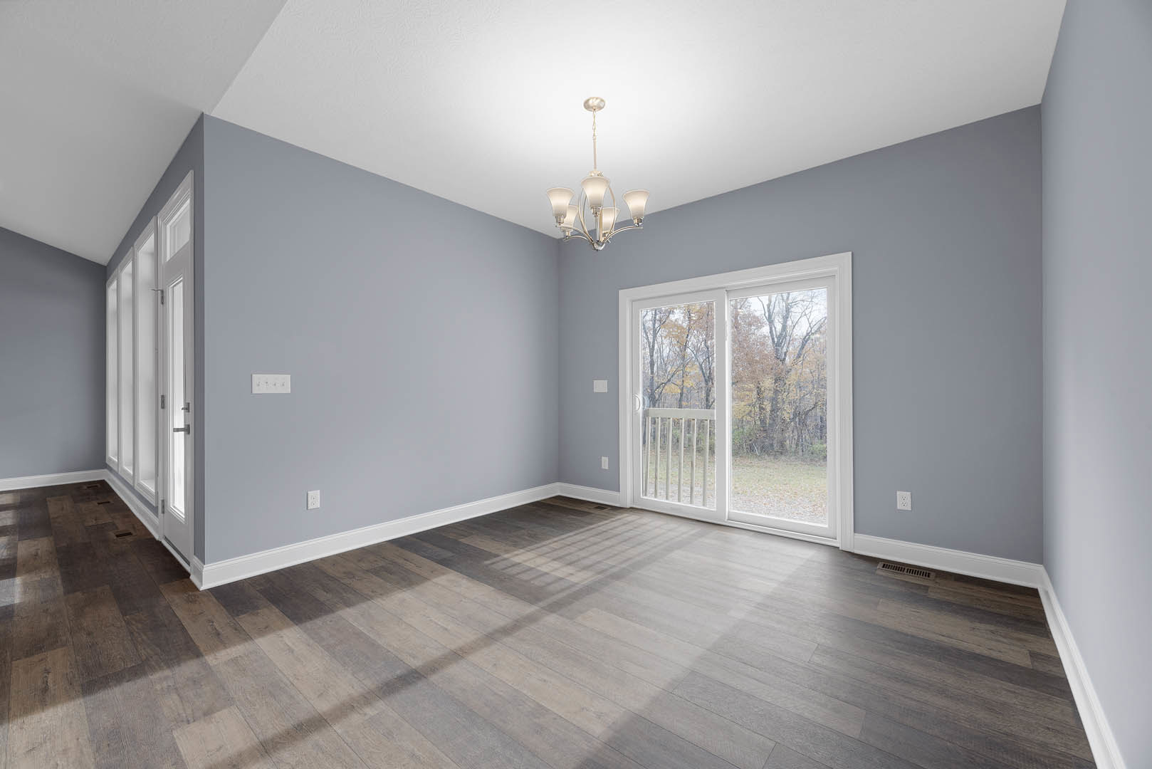 Open living space with wood flooring, sliding glass door overlooking trees, white walls, window with blinds, and modern chandelier hanging from ceiling