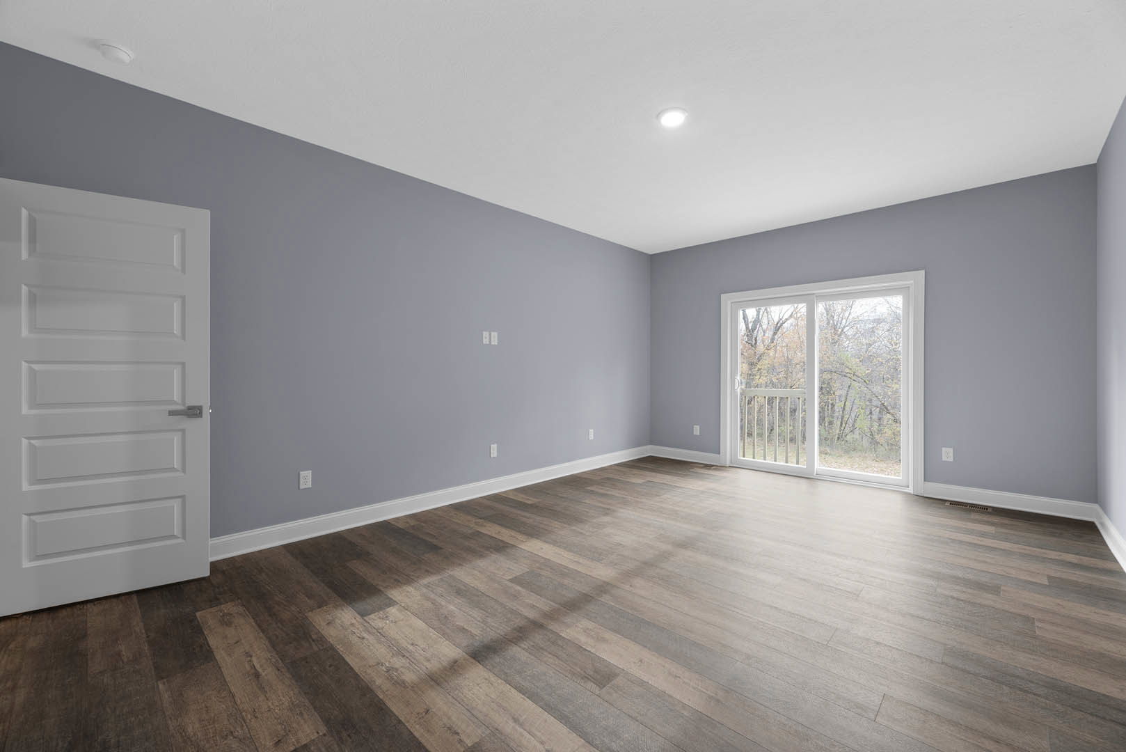 Wood flooring in a room with a white door featuring a silver handle, sliding glass door with white railing, plaster walls, and a window overlooking trees.