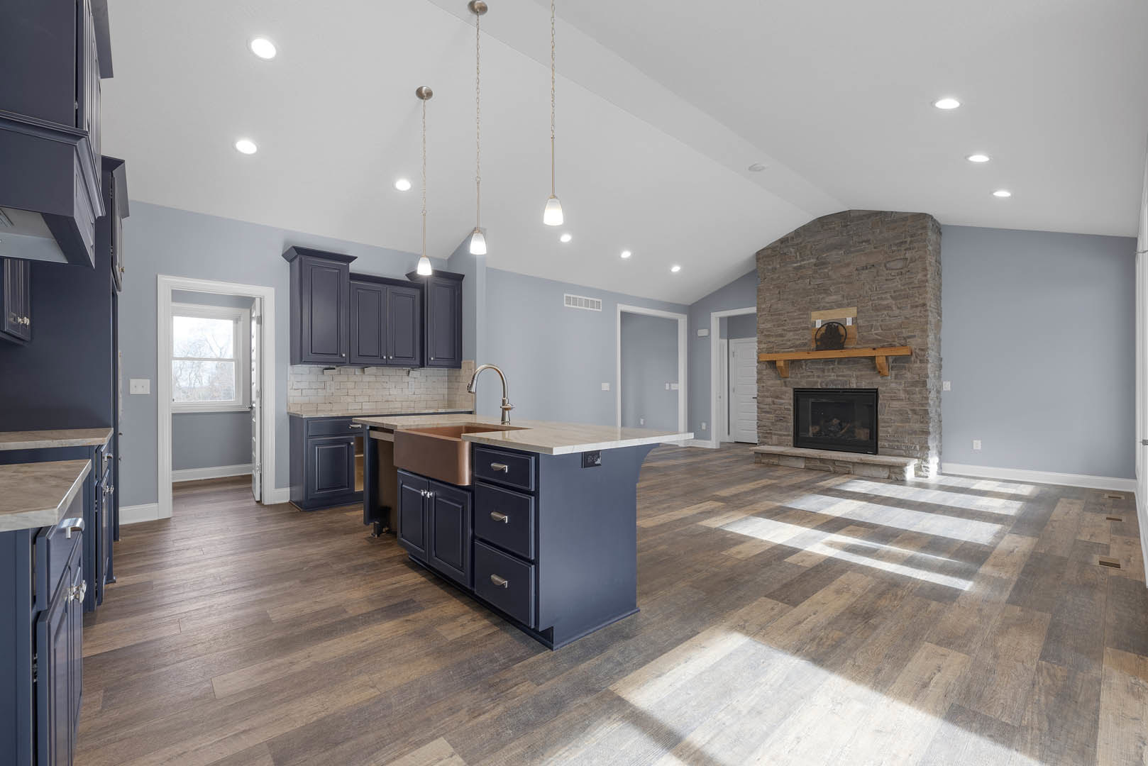 Kitchen with stone fireplace featuring glass door and wooden mantel, copper sink set in kitchen island with drawers, white-framed window, light cabinetry, and neutral flooring