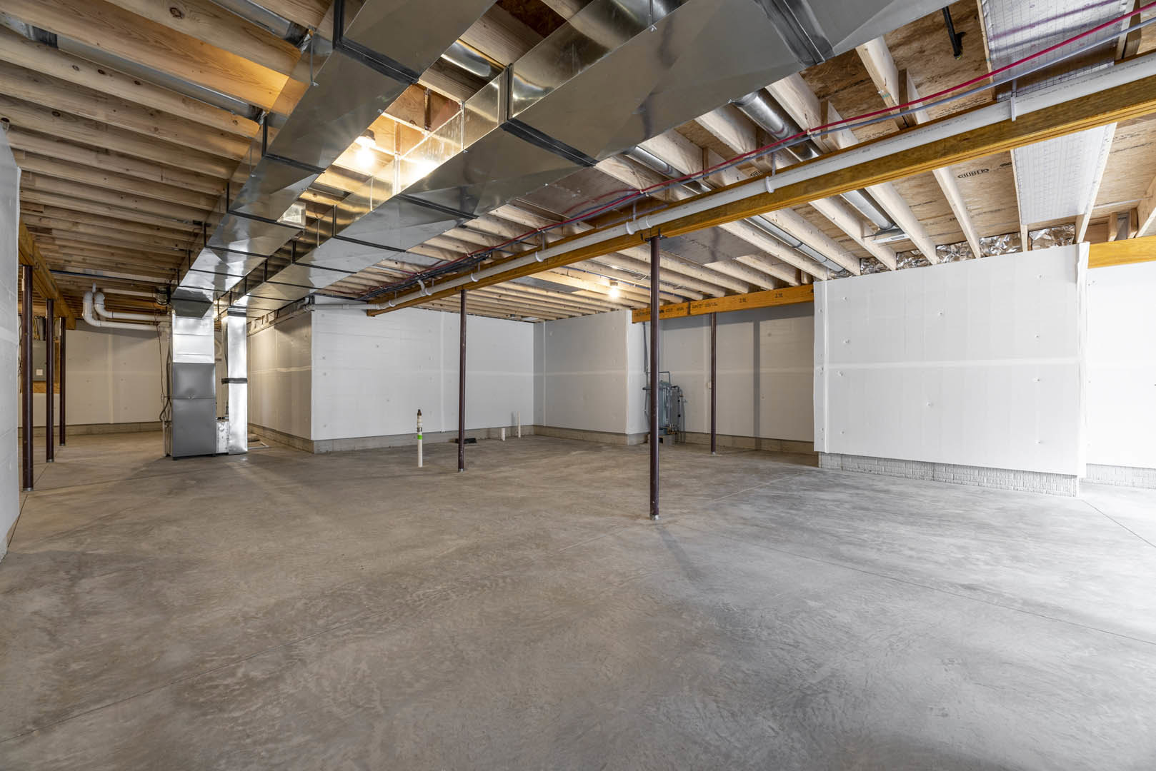 Exposed metal beam and metal ceiling above concrete floor with visible pipes, white wall with metal studs, and wooden pole with metal frame in unfinished room
