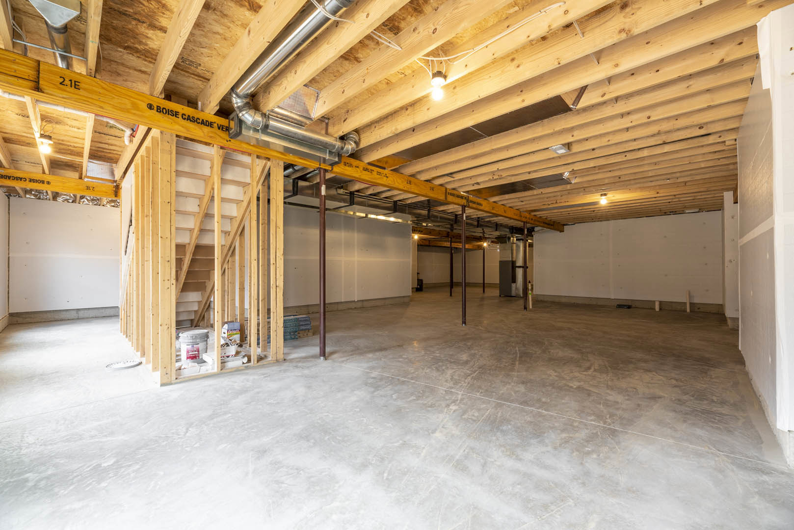 Room with exposed wooden ceiling and central beam, unfinished concrete floor, metal support poles, stack of blue boxes, white paint bucket, partially insulated walls