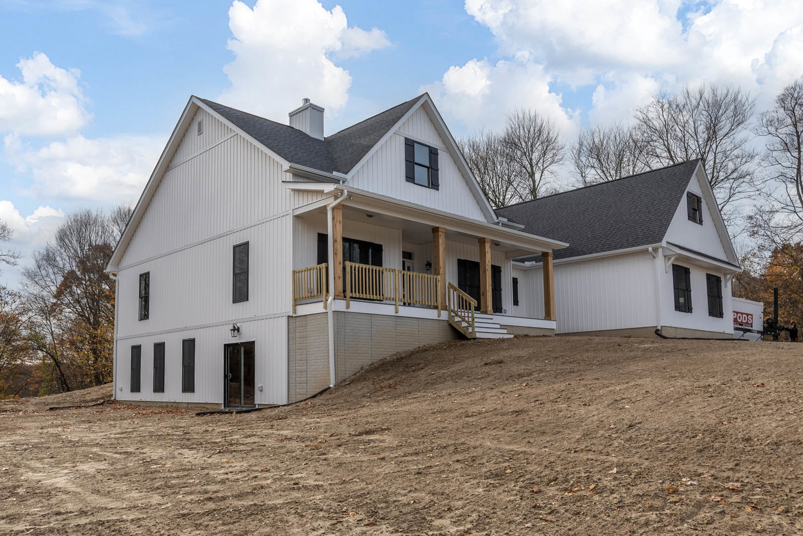 Two-story cottage with white siding, front porch and stairs, large windows, surrounded by trees, dirt hill in foreground, Robert Frost Farm visible in background under cloudy sky