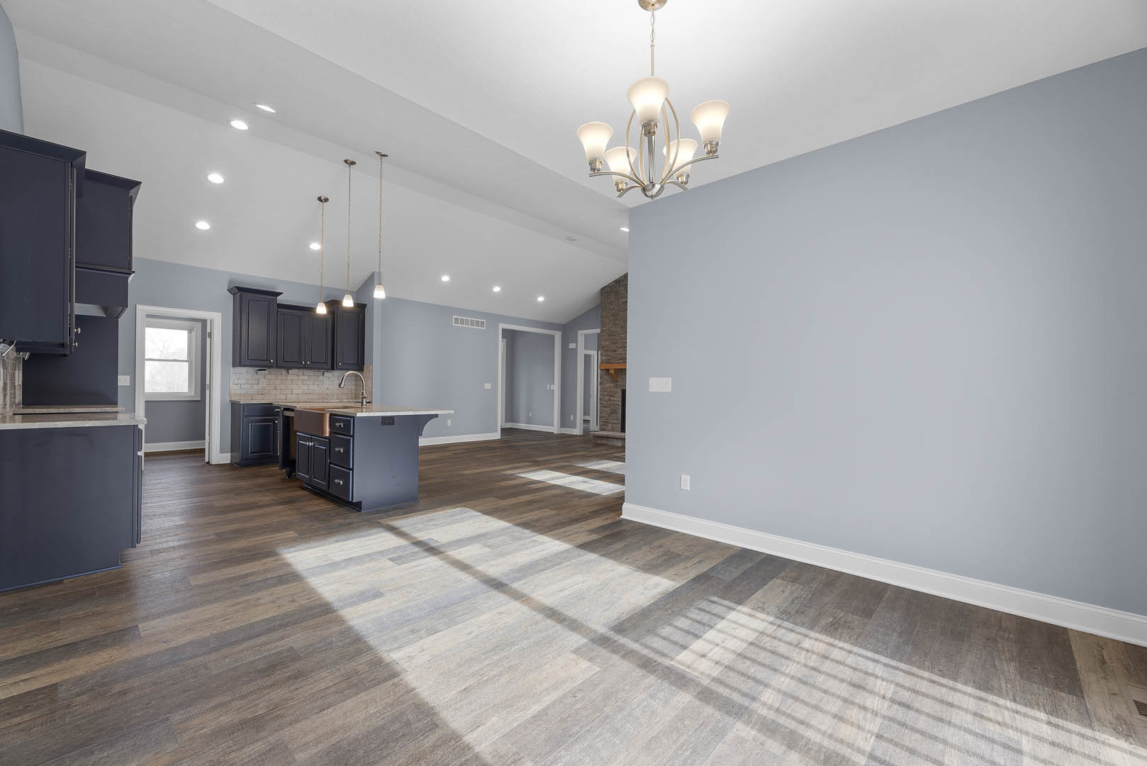 Open-concept kitchen with wood flooring, white cabinetry, large central island with drawers, chandelier overhead, sunlight streaming through windows, and adjacent white door with