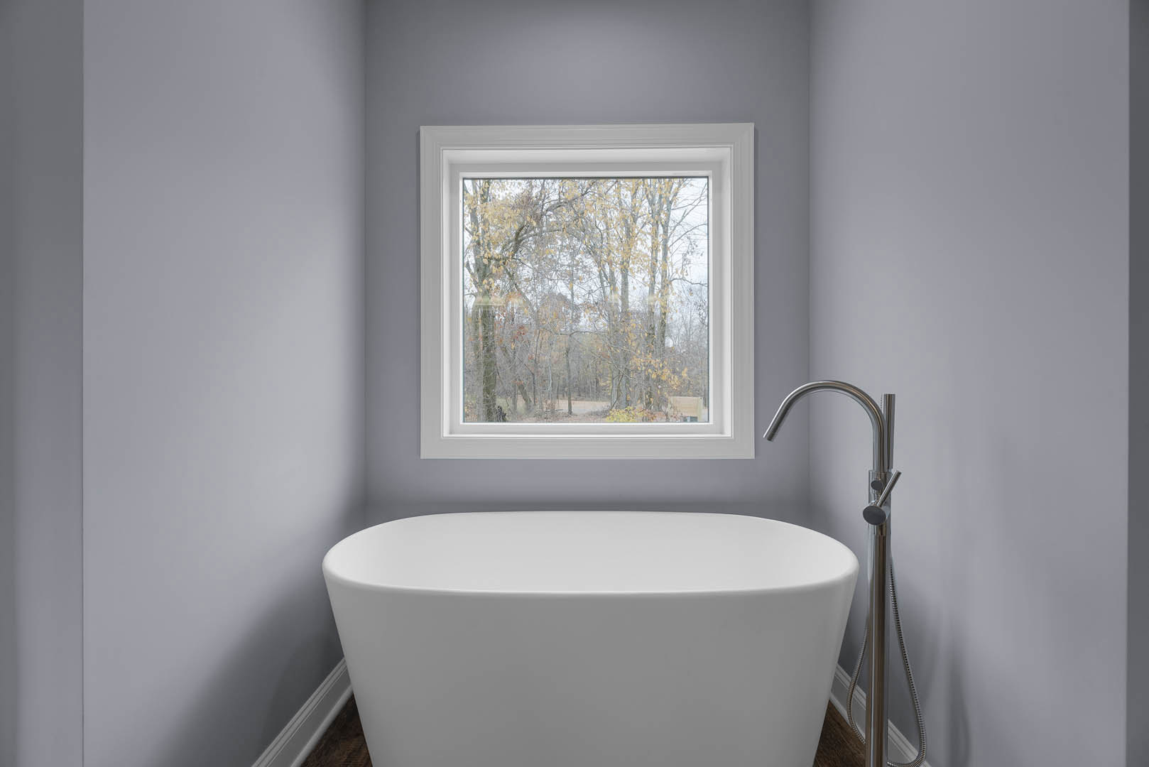 Freestanding white bathtub beside large window with outdoor tree view, light grey walls, chrome faucet, and nearby white sink