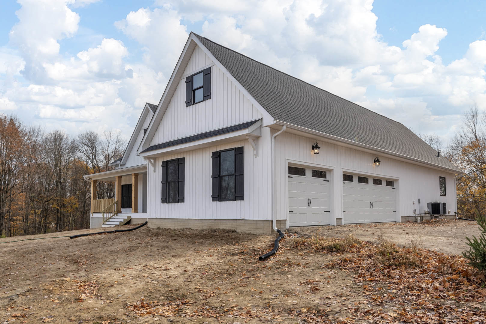 White siding house with attached garage, black window shutters, black downspout, and mature trees in the background under a cloudy sky