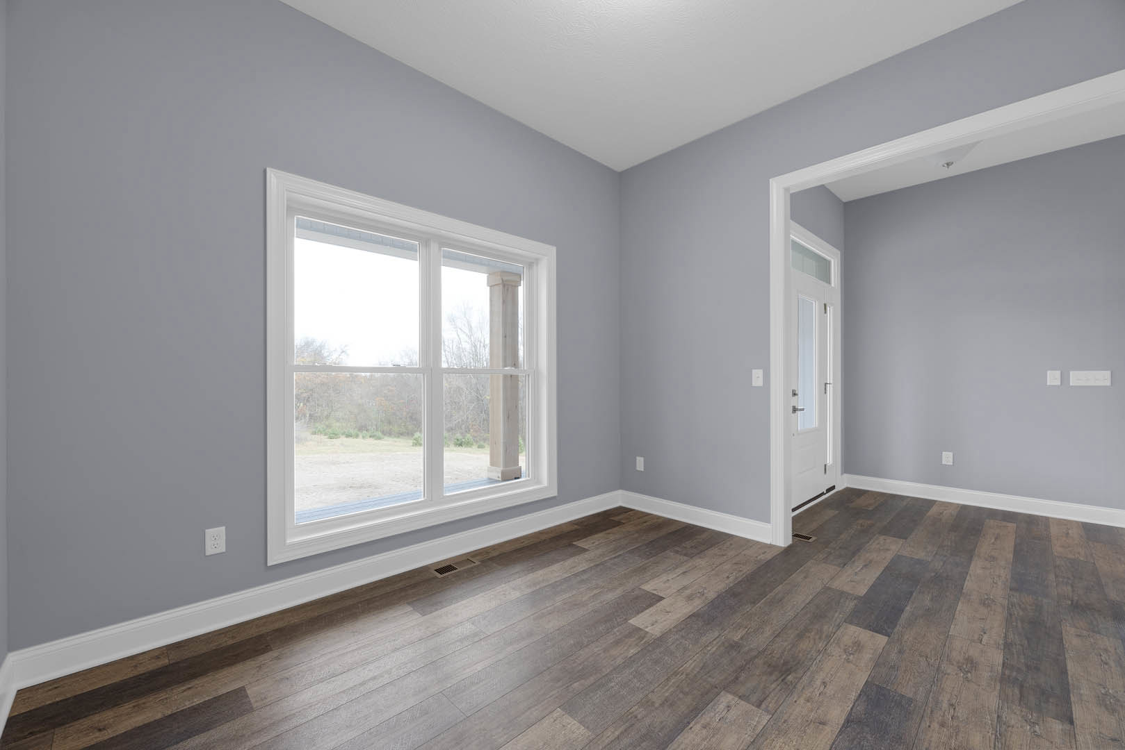 Sunlit room featuring wide-plank wood flooring, white walls, and a large window with a white frame overlooking a green yard.