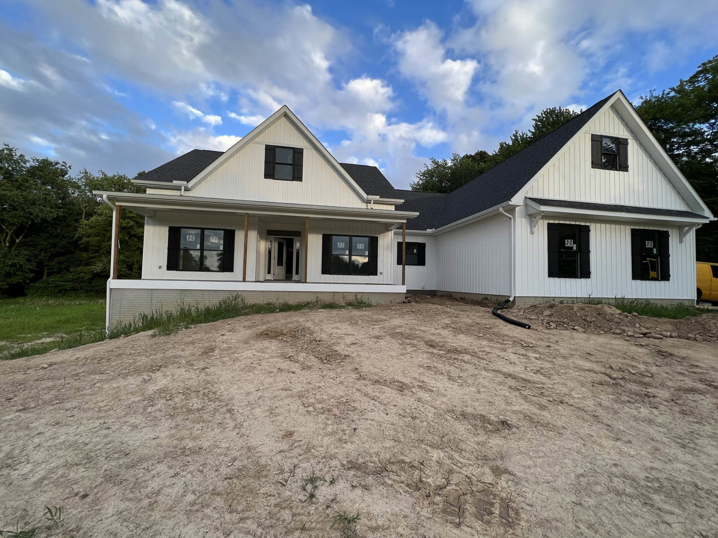 White custom home with covered porch, black shutters, and large windows, set against a dirt hill and open field under a blue sky