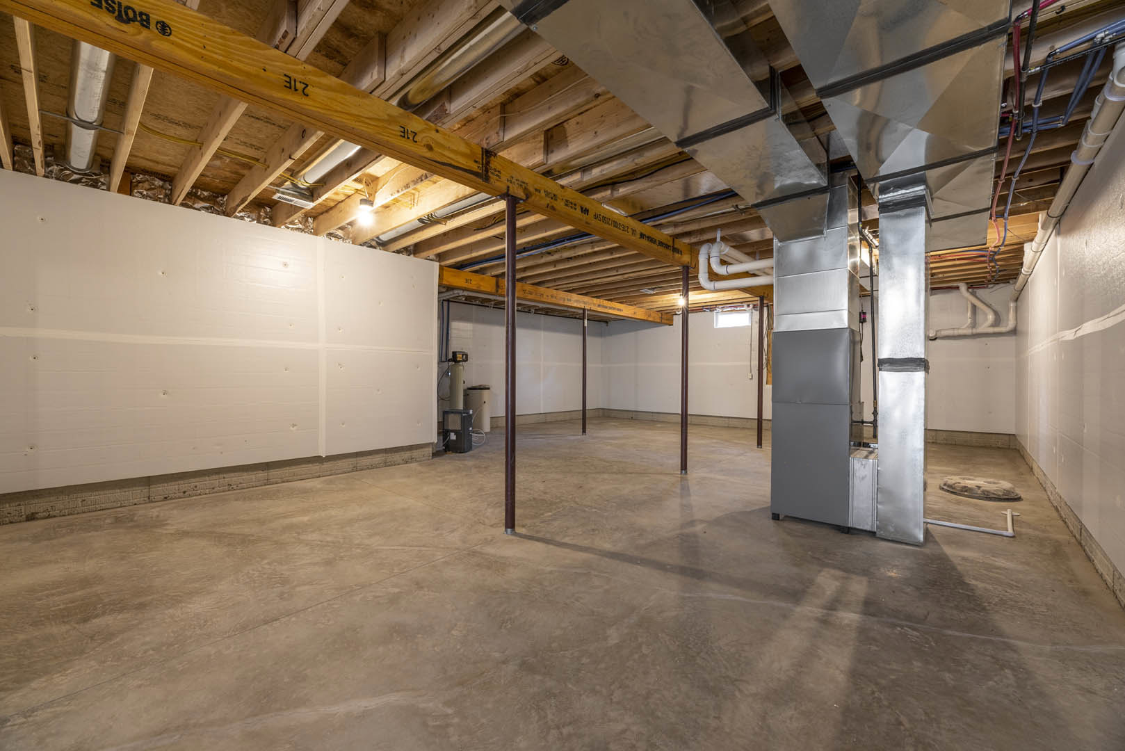 Exposed metal pipes and wood ceiling beams above a concrete floor, white wall and column with black electrical box, striped white towel hanging nearby in unfinished basement space