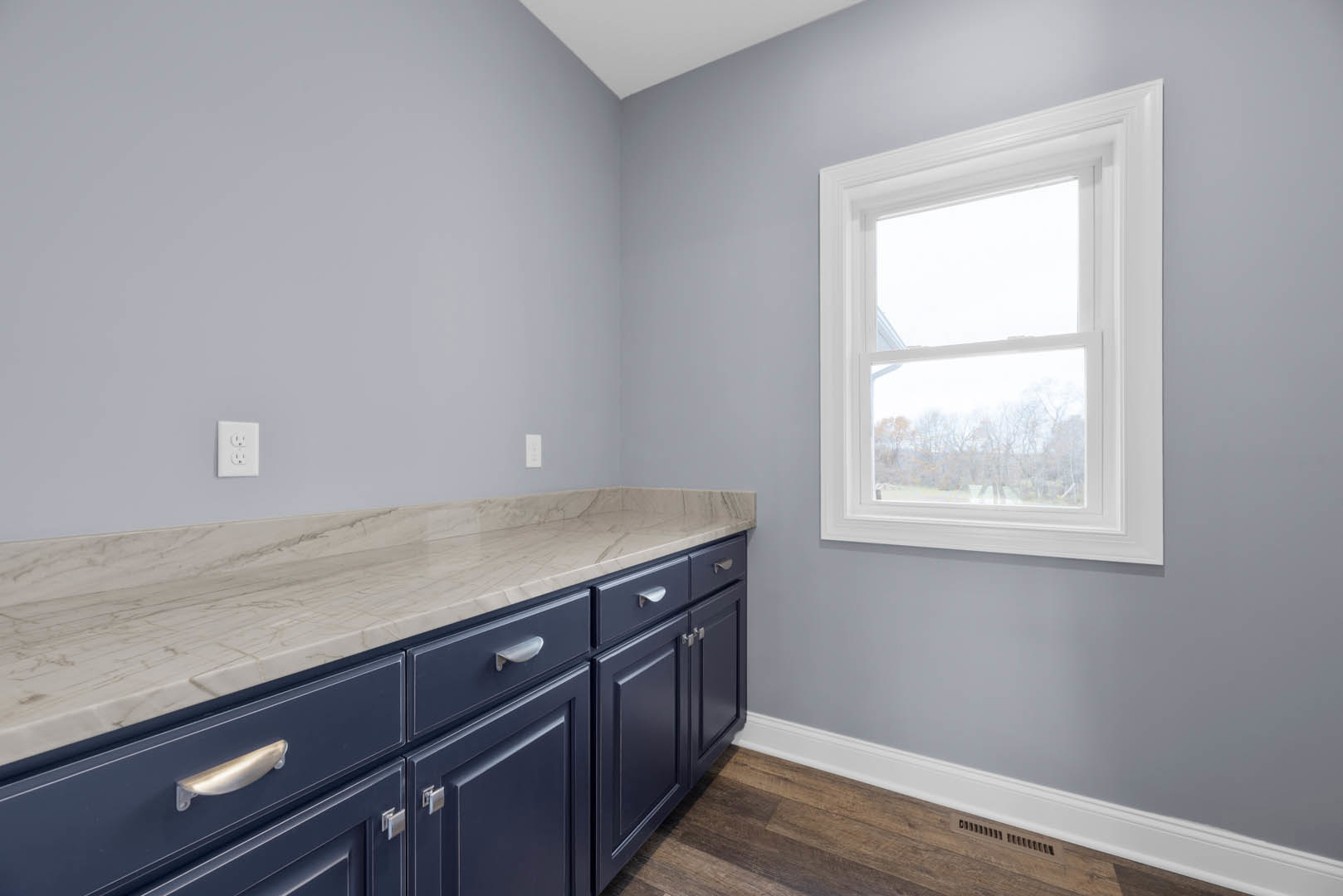 Kitchen with white marble countertop, stainless steel sink, light wood cabinetry, drawers, and large window overlooking trees