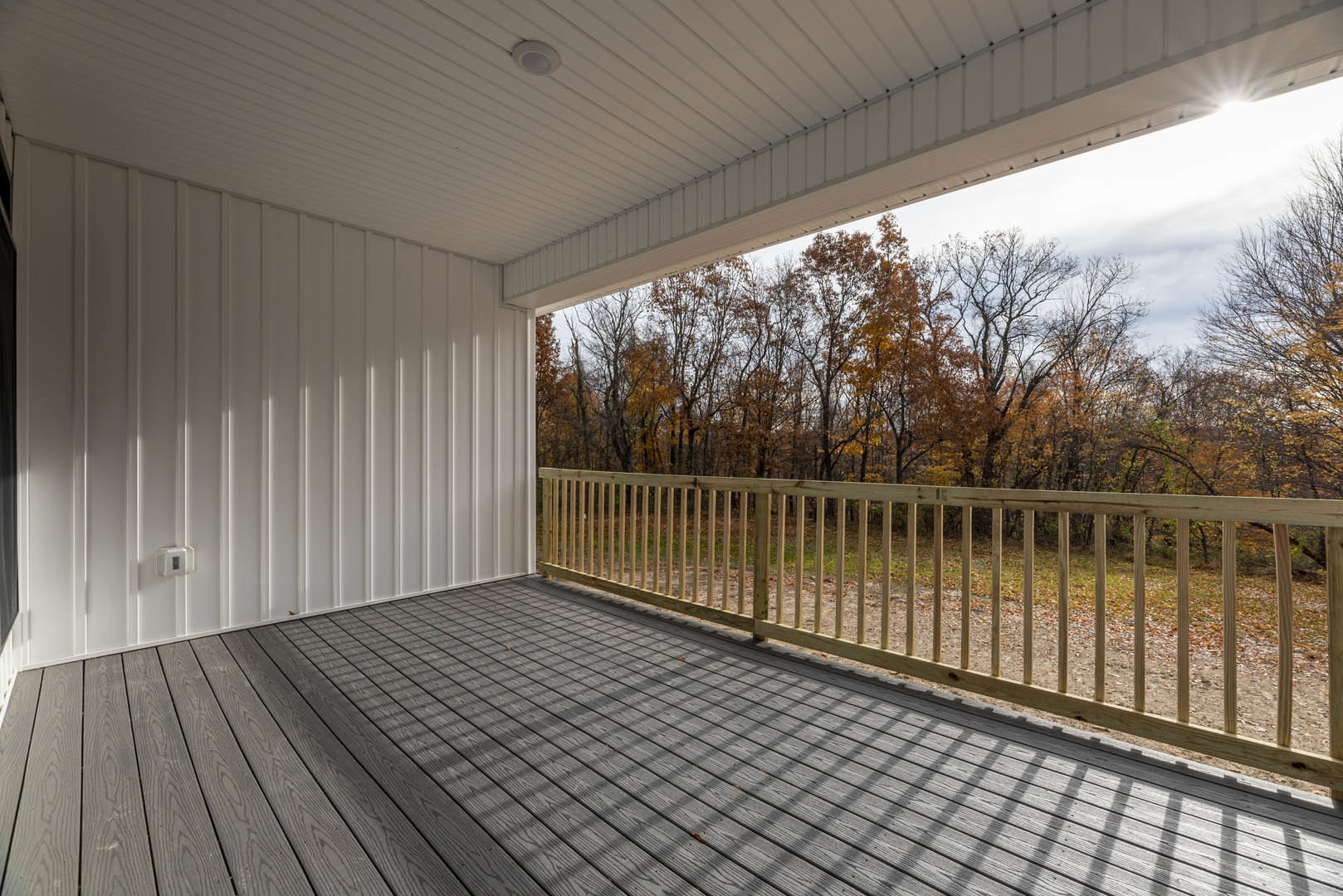 Wooden deck with metal railing overlooking leafy trees, white ceiling with recessed light, tile flooring, large window, and white walls.