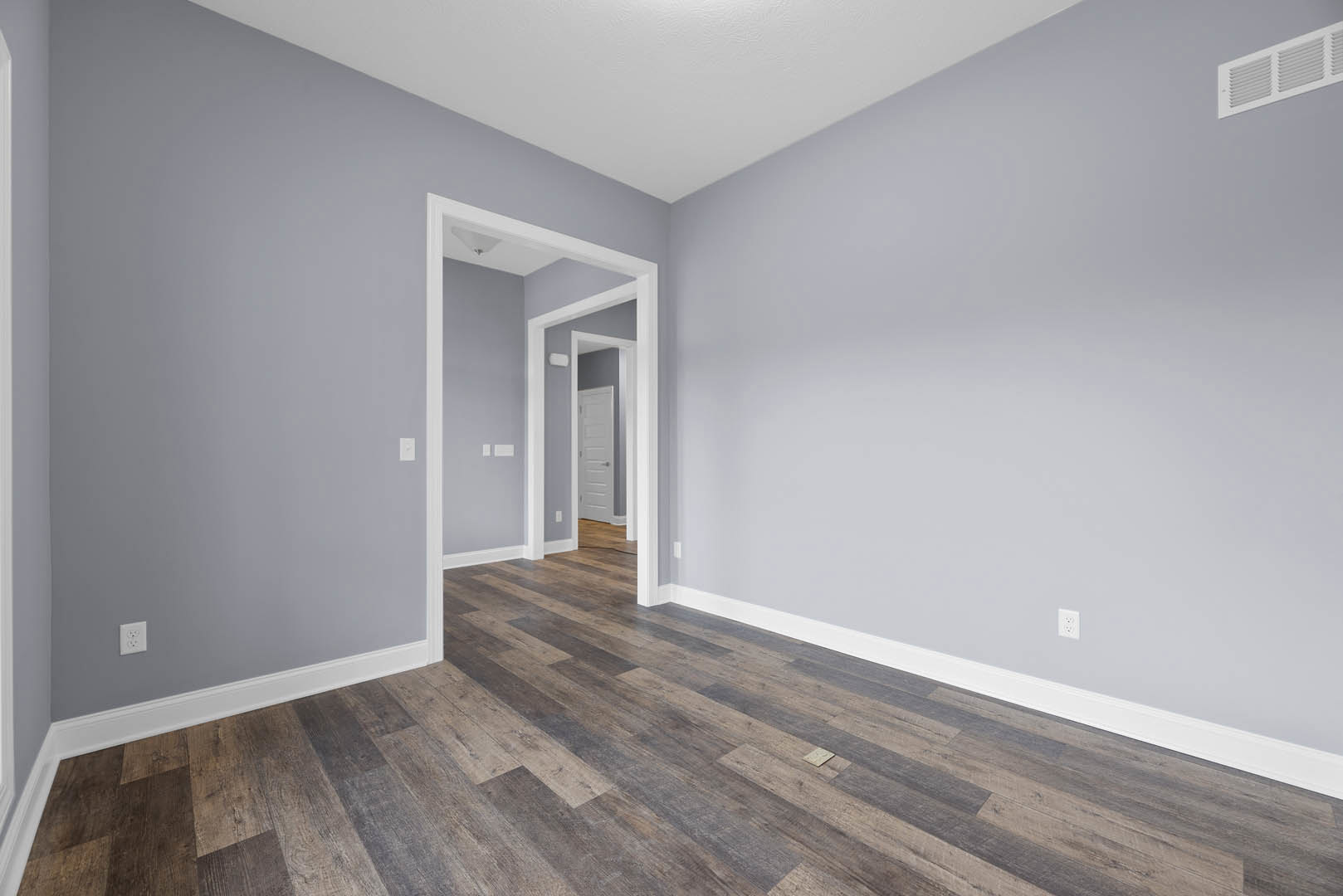 Wood flooring in a room with white walls, a white door, light switch, ceiling corner, and wall vent