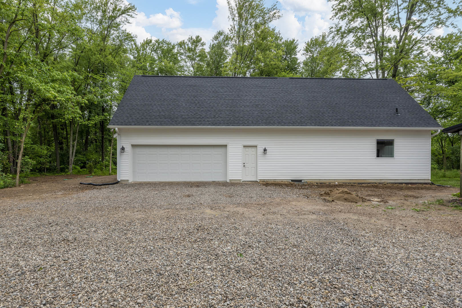 White house with black roof, white garage door with white trim, gravel driveway, white siding, trees in background