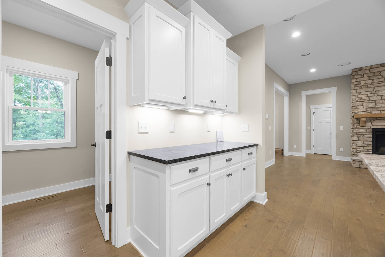 White kitchen cabinets with black countertops, wood flooring, a window overlooking trees, white door, and sink area.