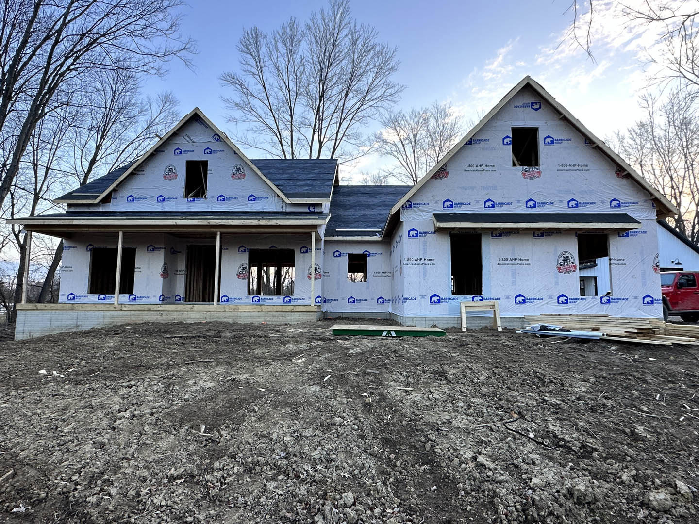 Partially built house with exposed framing, blue construction wrap, dirt yard, and mature trees in the background