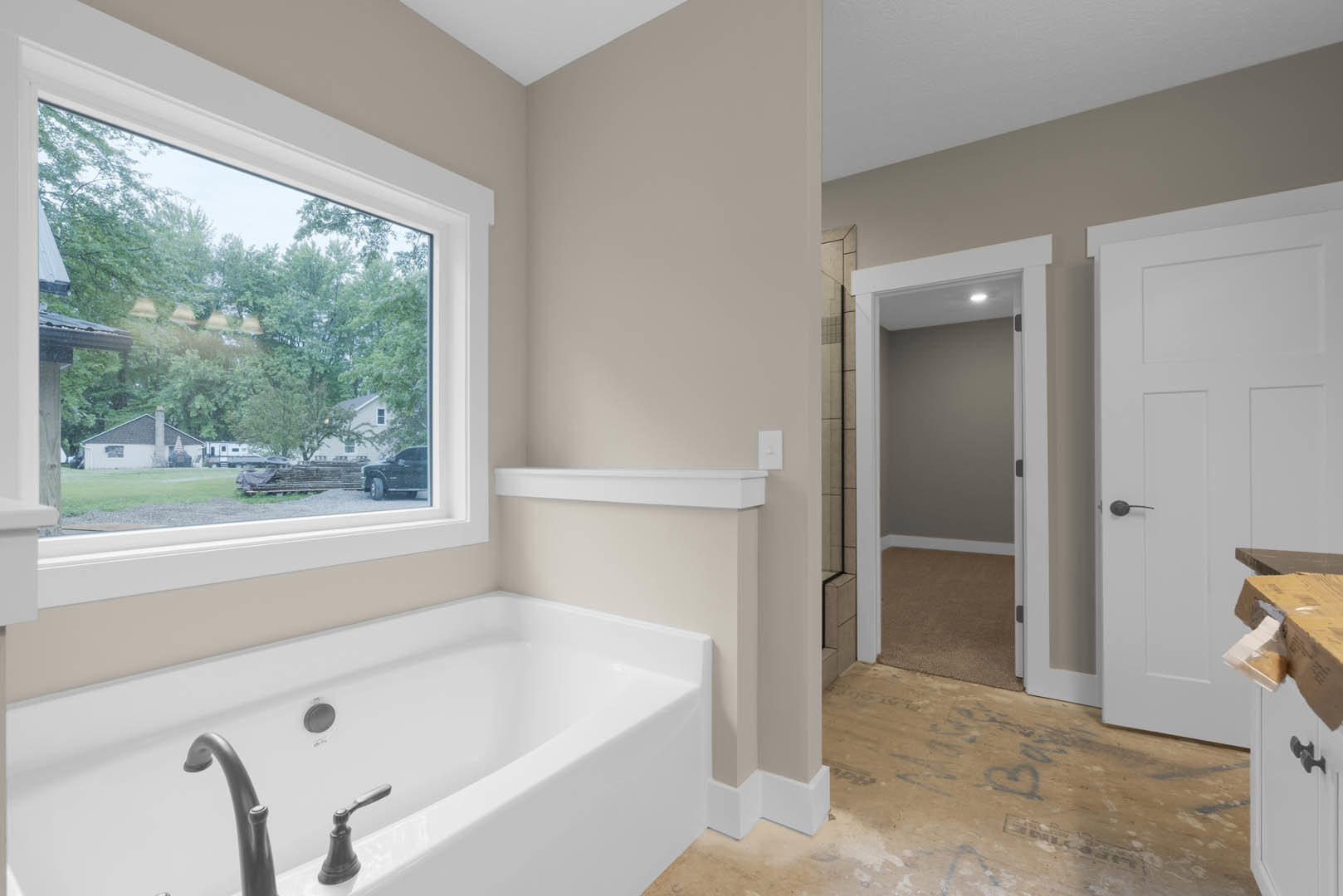 Freestanding white bathtub with chrome faucet beneath large window, light gray tile floor, white walls, and white door with black handle