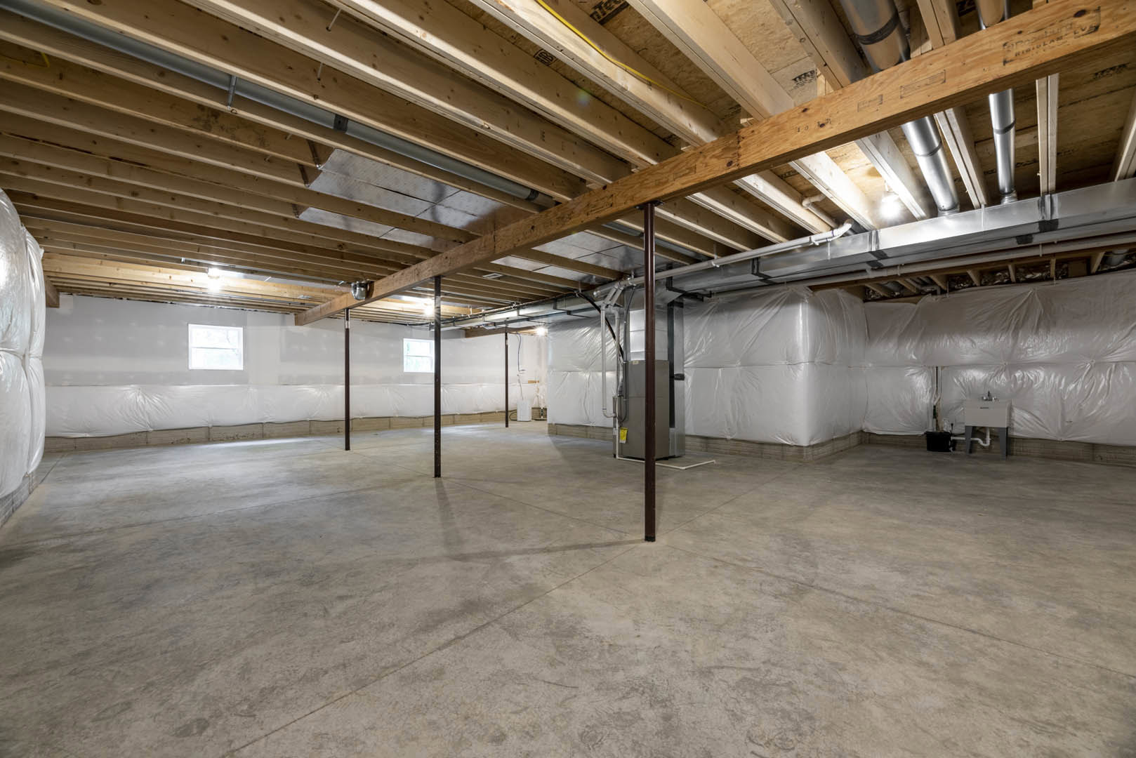 Concrete floor with exposed wooden ceiling beams, white walls, large window with white frame, and white plastic coverings scattered throughout the unfinished basement room.