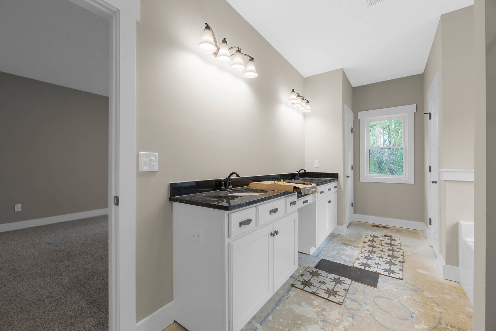 White kitchen cabinets with black granite countertops, stainless steel sink, tile backsplash, window overlooking trees, row of wall-mounted lights, white light switch, carpeted